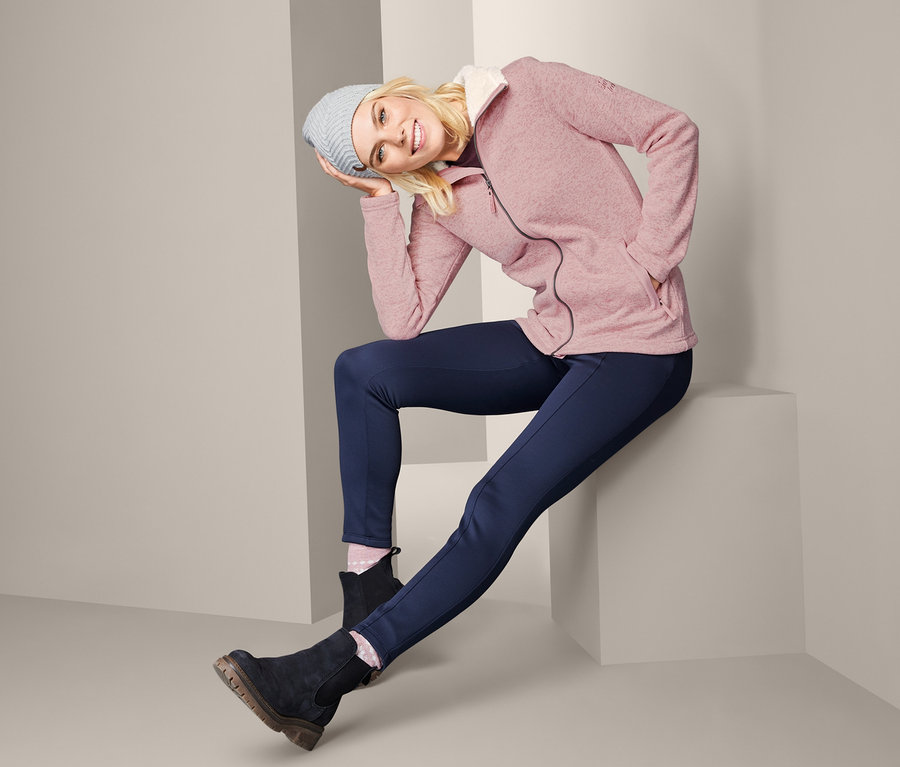 Blonde Frau sitzt und trägt eine Strickfleecejacke, blaue Hosen, Stiefel und Mütze.