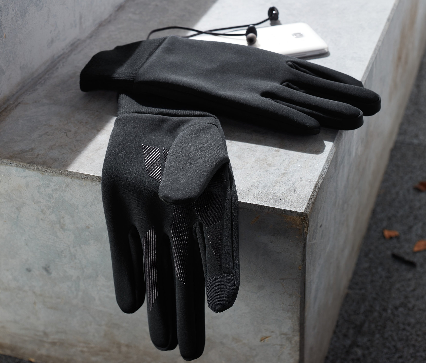 Zwei schwarze Windprotection-Handschuhe liegen auf einer Betonbank neben einem weißen Handy und Kopfhörern.
