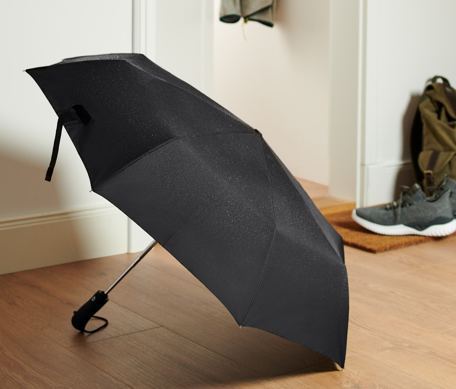 Ein schwarzer Automatik-Taschenschirm liegt auf einem Holzboden vor einer Tür.