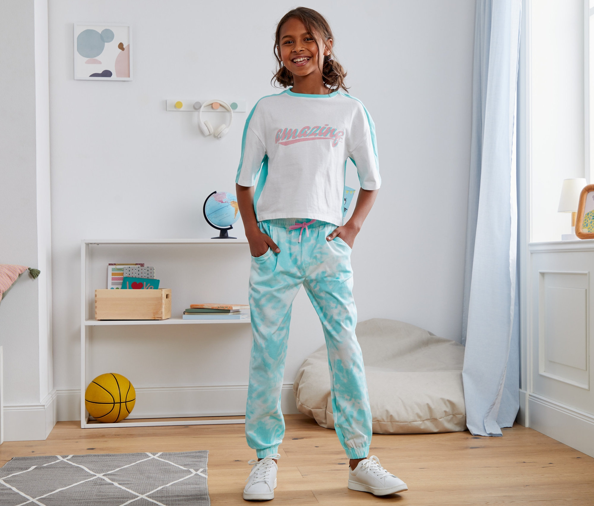 Lächelndes junges Mädchen steht im Kinderzimmer und trägt eine Jersey-Schlupfhose aus Bio-Baumwolle und ein T-Shirt.