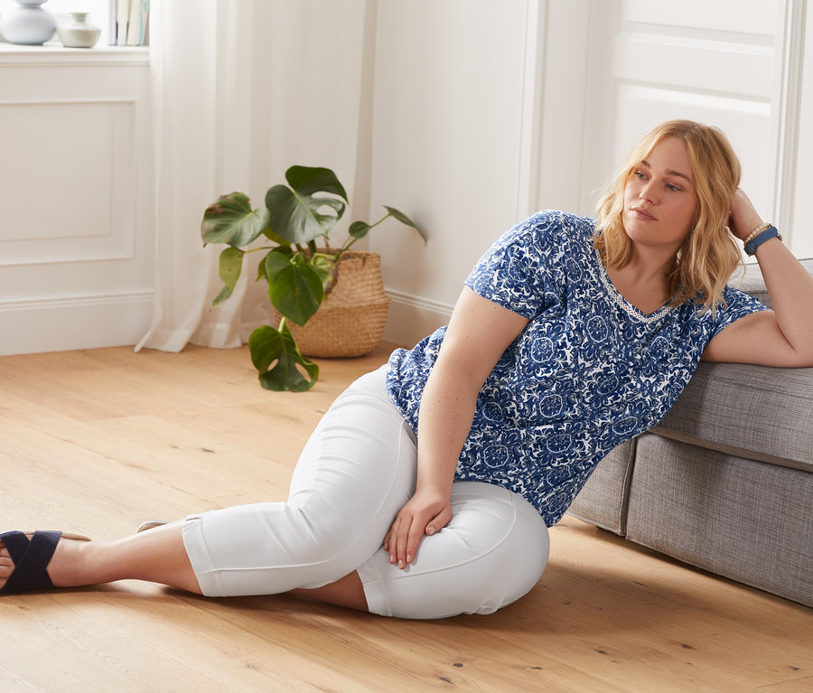 Eine Frau liegt auf dem Boden und lehnt sich an ein Sofa. Sie trägt eine weiße 7/8-Stretchhose und ein blaues, bedrucktes Blusenshirt.