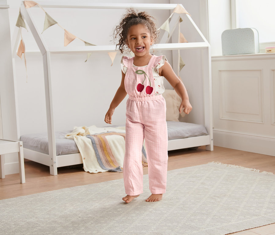Ein lächelndes kleines Mädchen trägt einen rosafarbenen Jumpsuit mit Leinen und Kirschen und steht in einem Kinderzimmer.