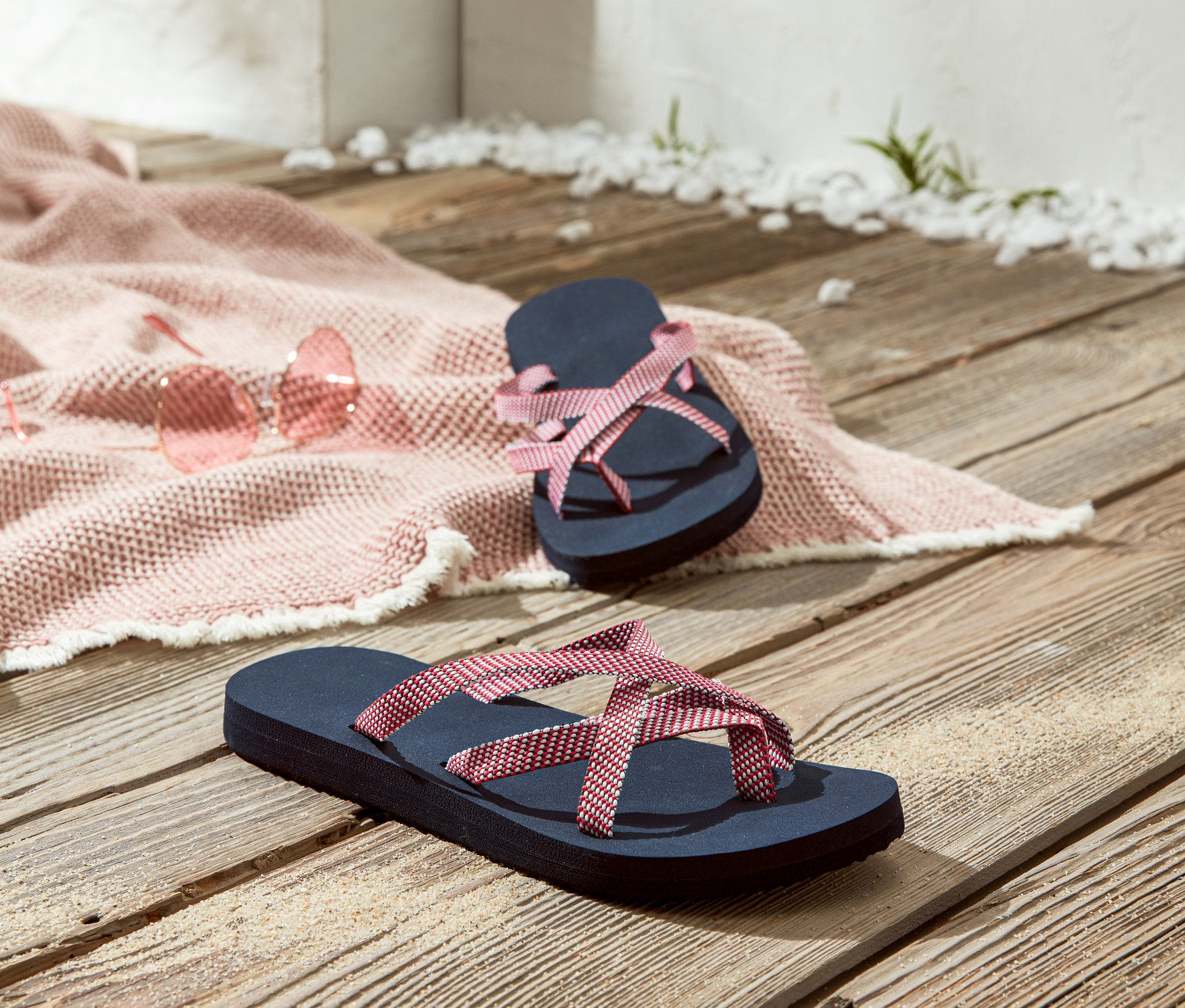 Zwei Zehengreifer liegen auf einem Holzboden neben einer rosafarbenen Sonnenbrille und einer rosa Stranddecke.