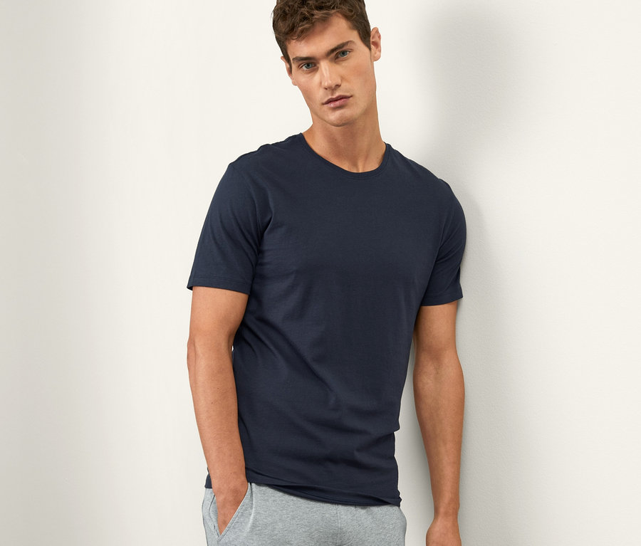 Mann lehnt an Wand, trägt blaues T-Shirt und graue Jogginghose. Er trägt 1 von 2 T-Shirts mit Rundhalsausschnitt.