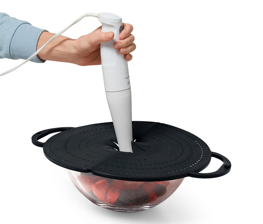 Eine Hand hält einen Stabmixer über eine Schüssel mit Früchten, die mit einem schwarzen 4-in-1-Pfannen-Spritzschutz abgedeckt ist.