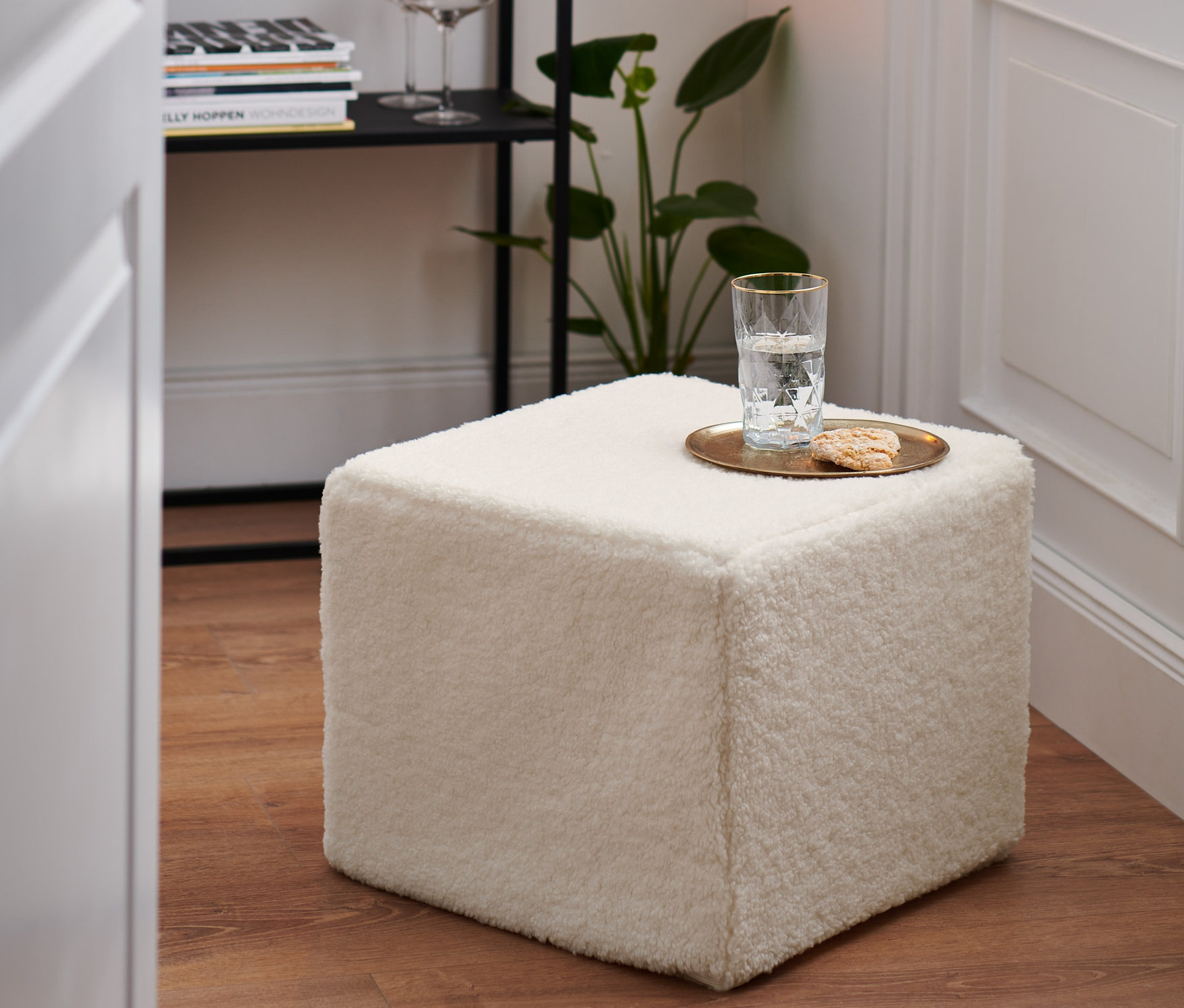 Ein cremefarbener Sitzpouf steht auf einem Holzboden. Auf dem Sitzpouf stehen ein Glas Wasser und ein Keks.