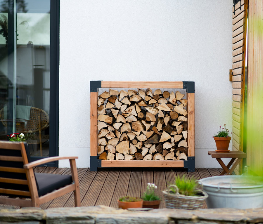 Ein Kaminholzständer, L, steht auf einer Terrasse neben einer weißen Wand.