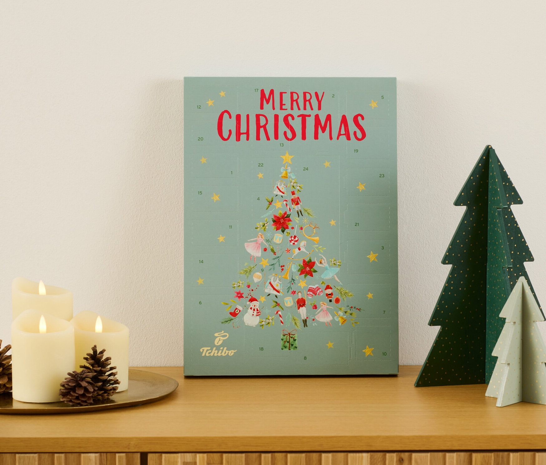 Tchibo Adventskalender auf einer Holzkommode neben Kerzen und Deko-Tannenbäumen.