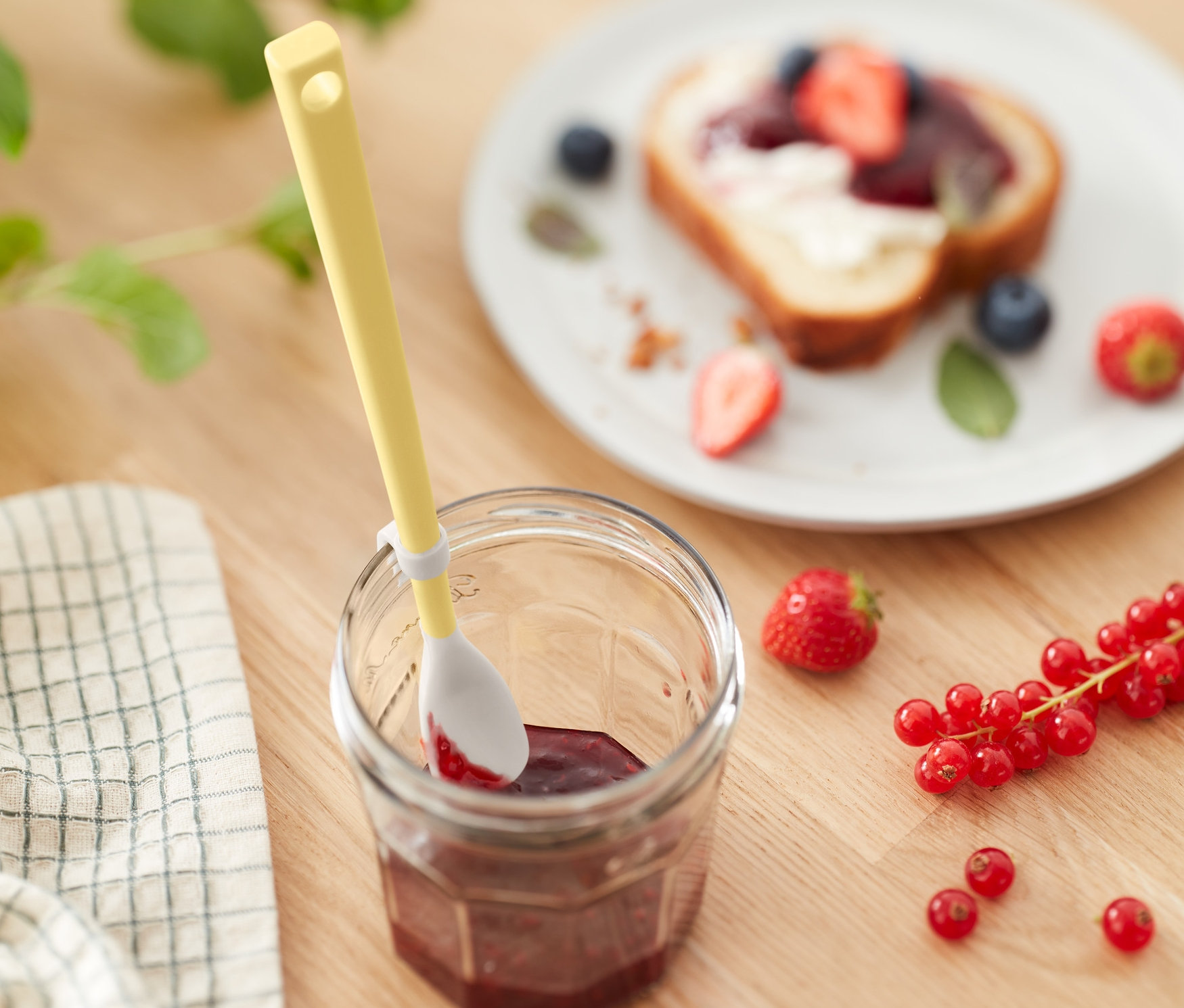 Ein gelber Spezial-Silikonlöffel steckt in einem Glas Marmelade, daneben Erdbeeren und Johannisbeeren, im Hintergrund ein Toast mit Früchten.