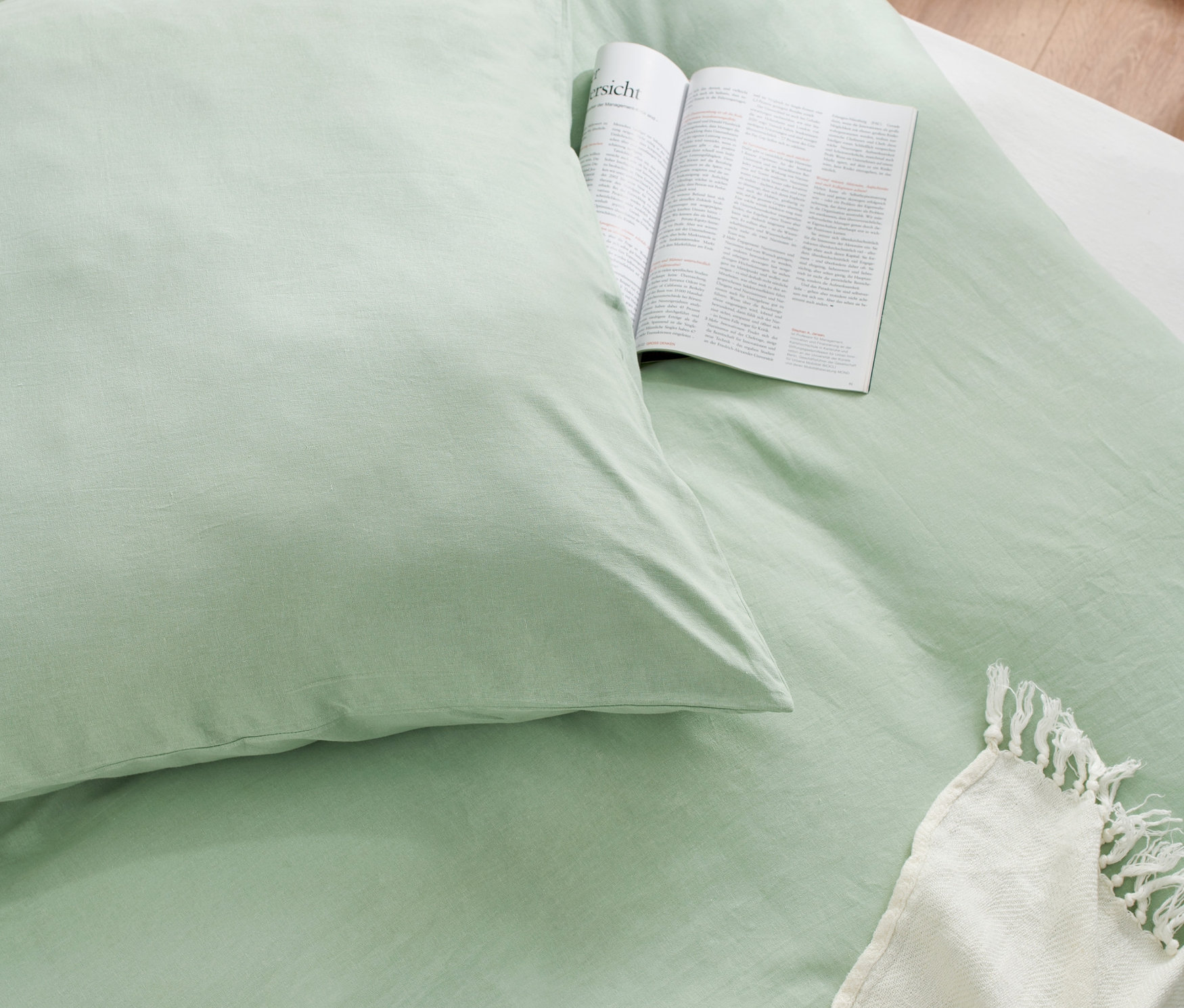 Nahaufnahme von hellgrüner Bettwäsche aus gewaschenem Leinen in Normal- und Übergröße mit offenem Buch und weißer Decke.