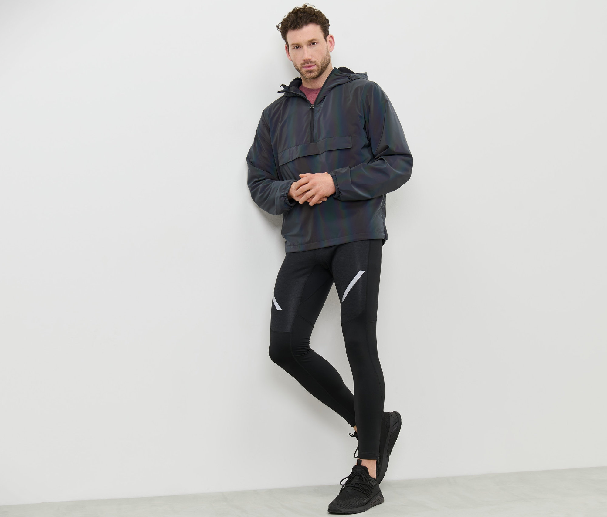 Mann lehnt an Wand, trägt Windbreaker Rainbow Reflective, schwarze Leggings und Turnschuhe.