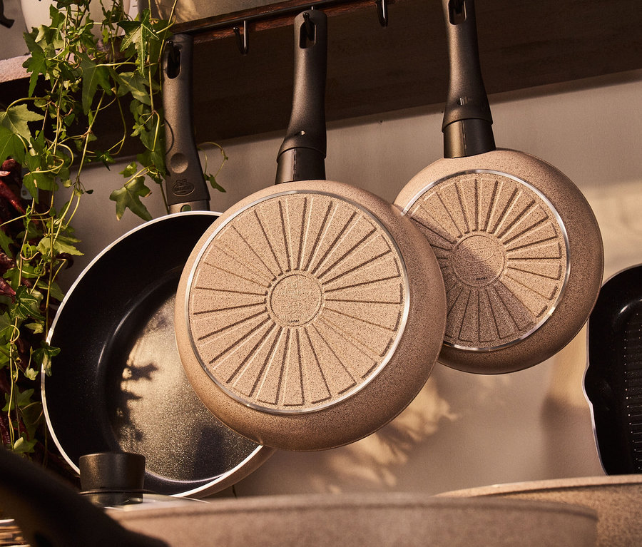 Drei sandfarbene BALLARINI Aluminium-Bratpfannen hängen an der Wand.
