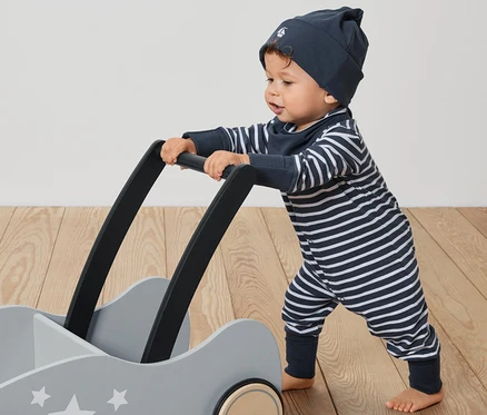 Kleinkind schiebt grauen Wagen mit Sternen. Es trägt ein Baby-Set, 3-teilig: gestreifter Overall, Mütze und Lätzchen.