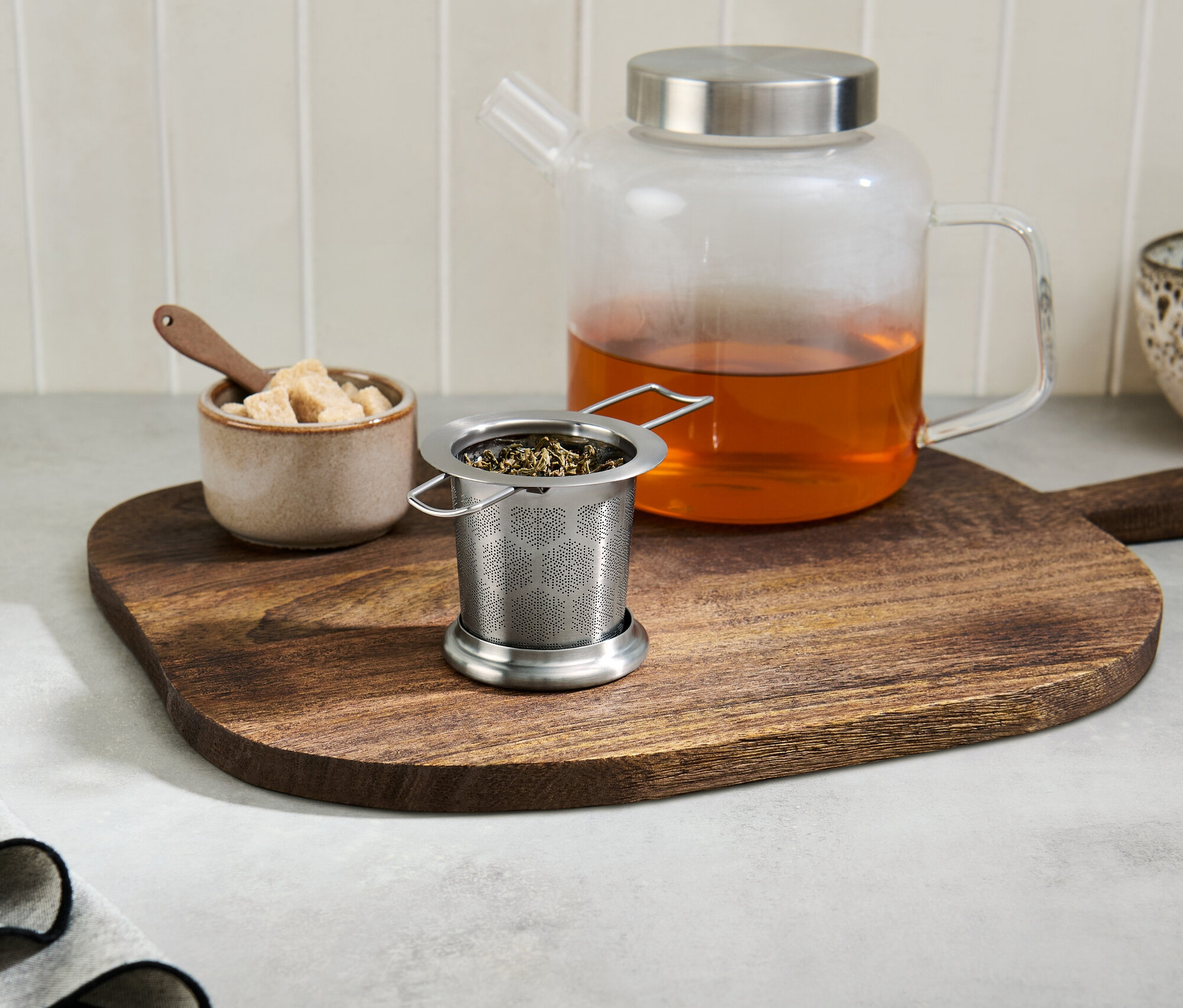 Teesieb mit Tee auf einem Holzbrett neben einer Glasteekanne mit Tee.