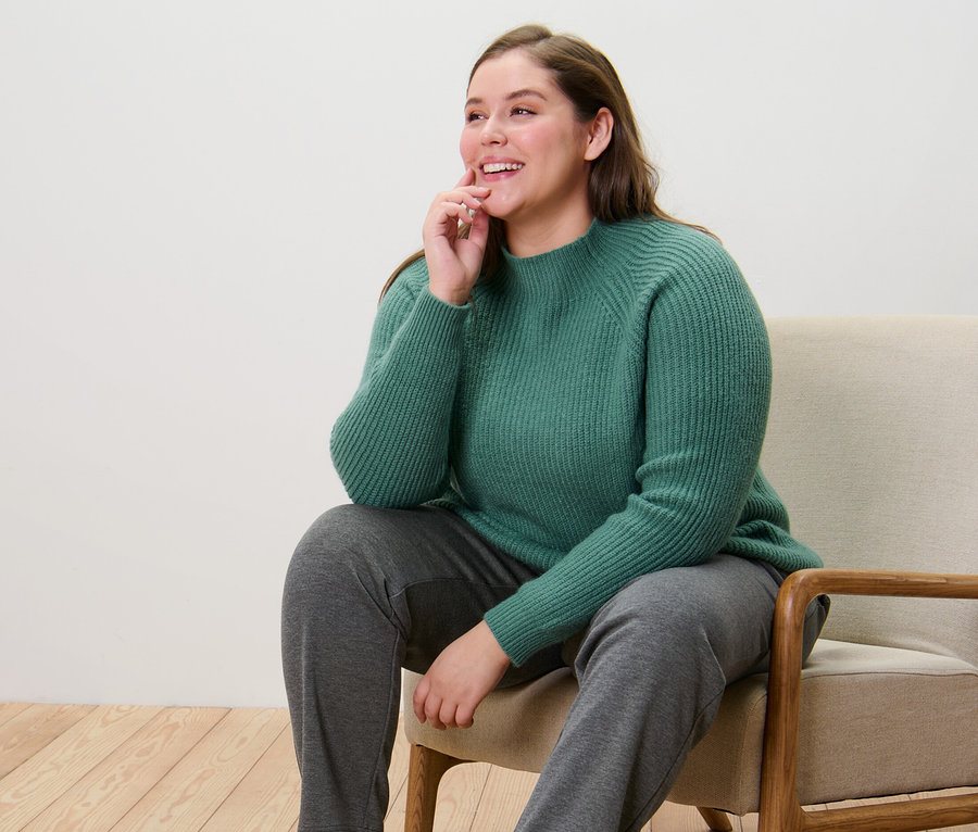 Eine Frau sitzt auf einem Stuhl und trägt einen grünen Grobstrickpullover mit Stehkragen und eine Joggpants.