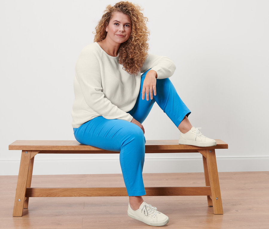 Frau sitzt auf einer Bank und trägt einen cremeweißen Strickpullover und eine Bengalin-Hose.