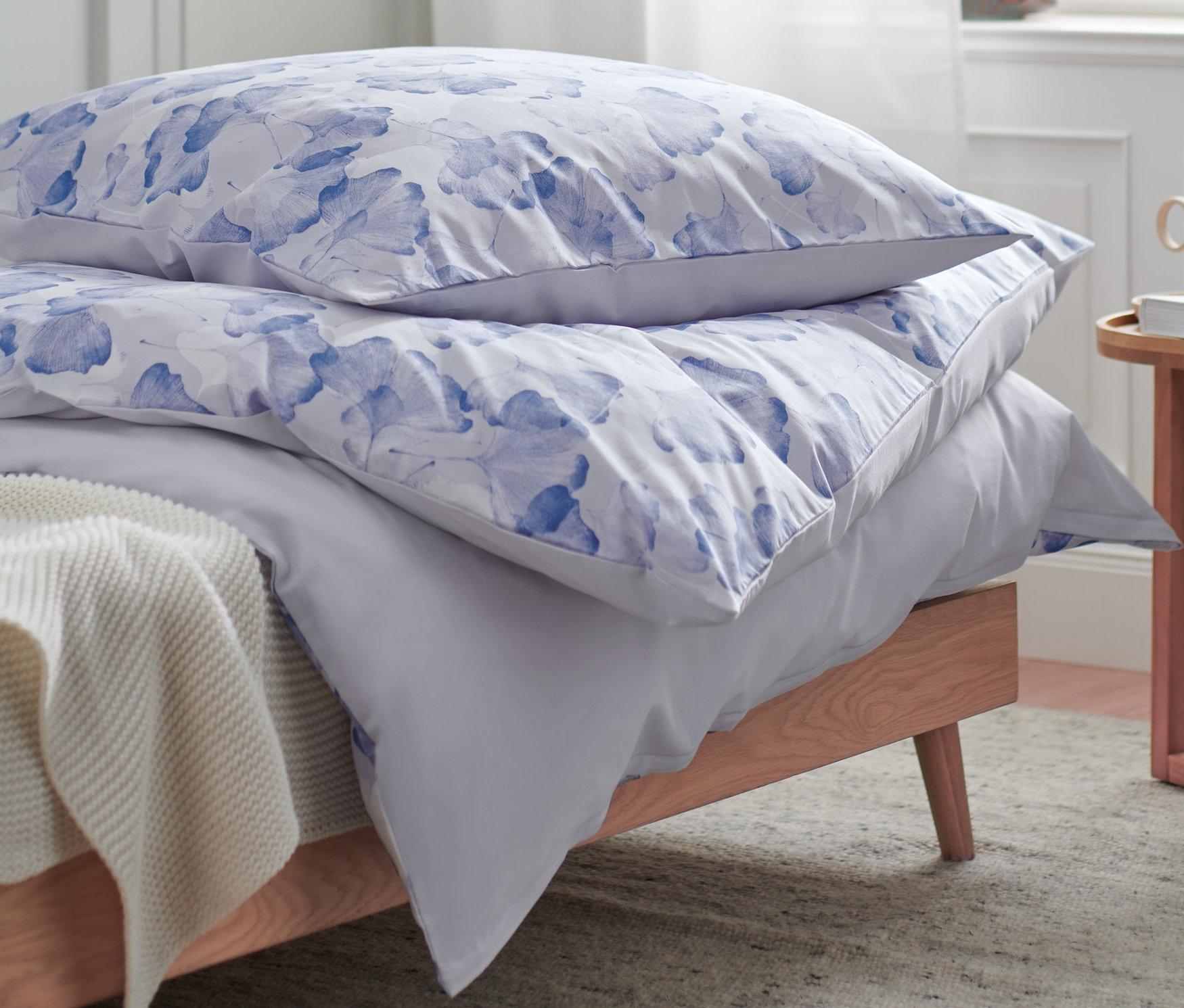 Zwei Kissen mit blauen Blumen liegen auf einem Bett mit grauer Bettwäsche neben einem kleinen Tisch.