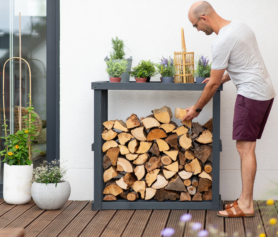 Mann legt Holzscheit in Kaminholzregal »Harald«. Auf dem Regal stehen Topfpflanzen.