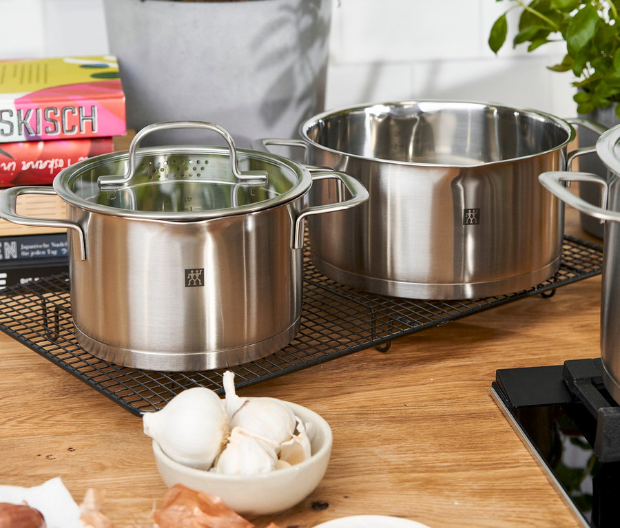 ZWILLING Trueflow Kochtopf-Set, 5-teilig aus Edelstahl auf einem schwarzen Gitter mit Zwiebeln und Knoblauch.