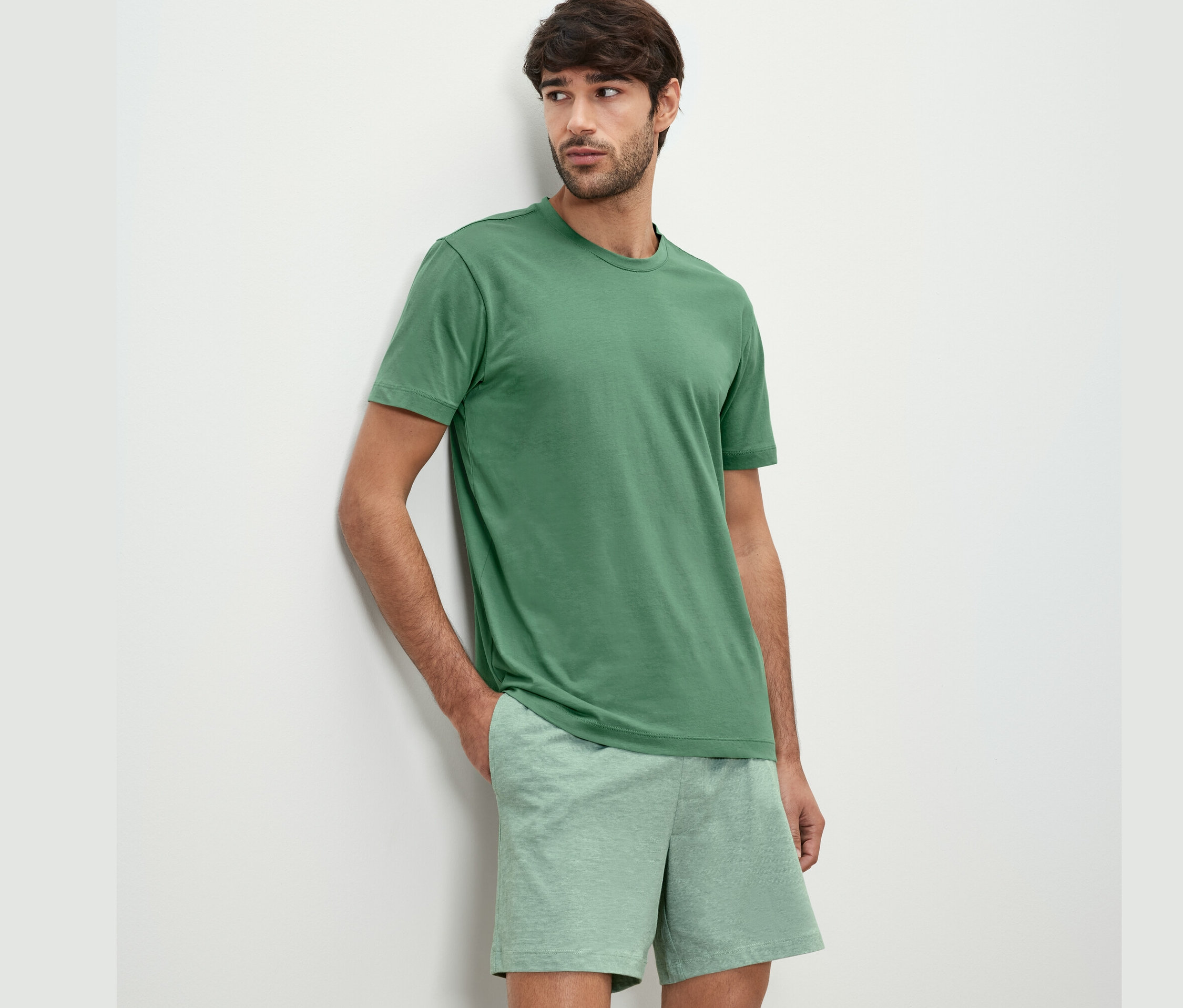 Mann lehnt an Wand, trägt Shorty-Pyjama-Set.