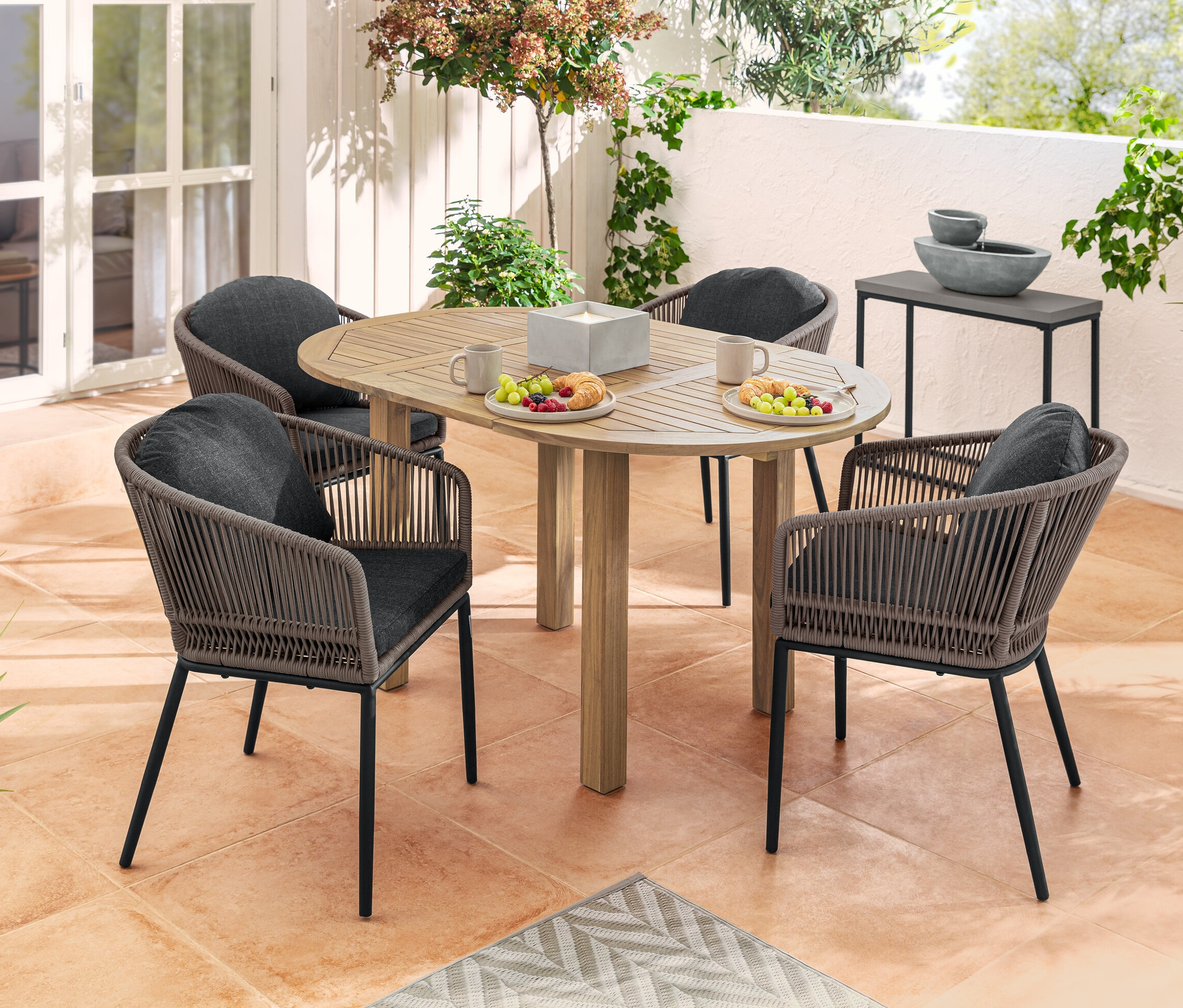 Vier Dining-Sessel mit runder Rückenlehne und Textilgeflecht um einen runden Ausziehtisch mit Speisen und Getränken auf einer Terrasse. Im Hintergrund Solar-Gartenbrunnen und ein Konsolentisch-Set.