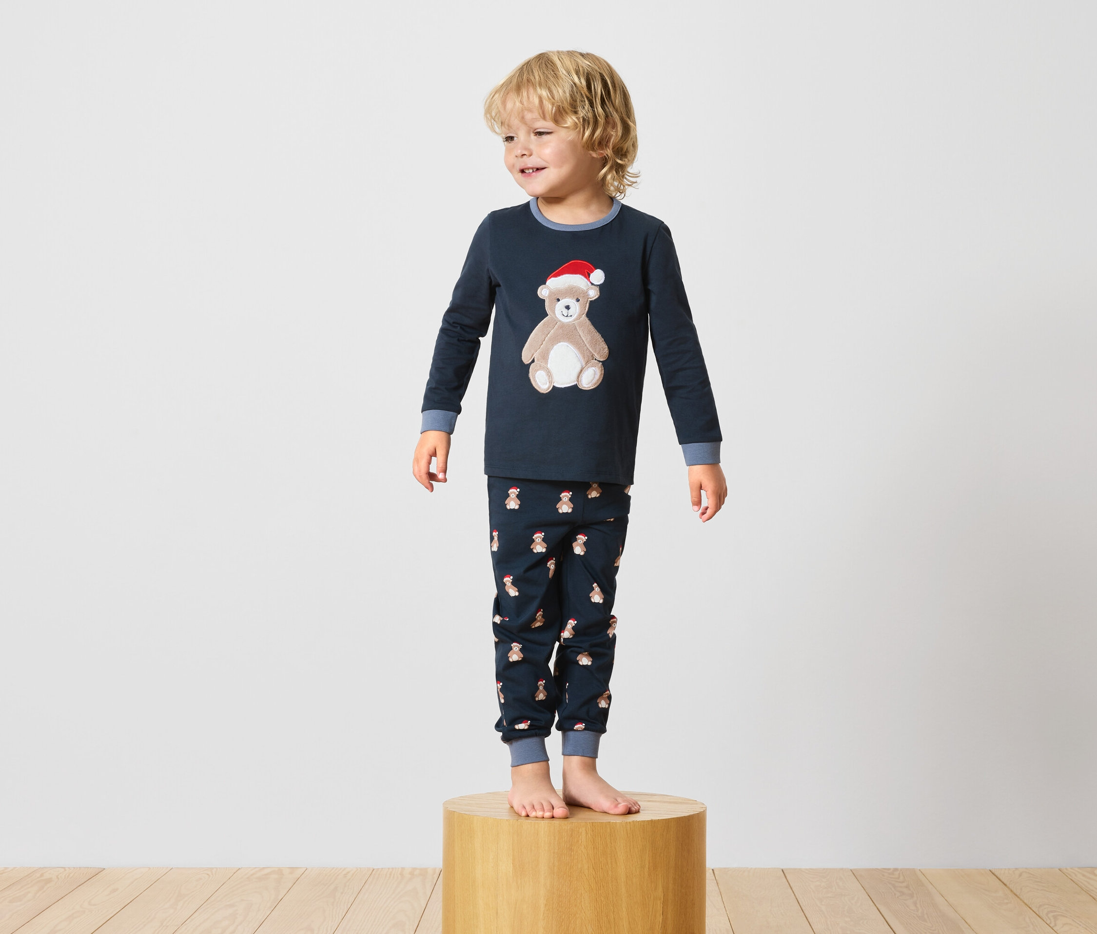 Junge steht auf einem Holzsockel und trägt einen dunkelblauen Pyjama mit einem Teddybär-Motiv.