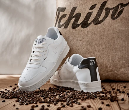 Zwei weiße Tchibo x VLACE Coffee Lover Sneaker liegen auf Kaffeebohnen vor einem Jutesack mit Tchibo-Logo.