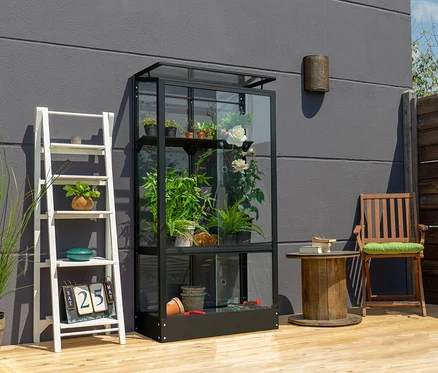 Ein schwarzes Vitavia Balkongewächshaus mit Standfundament steht auf einer Holzterrasse neben einem weißen Regal.
