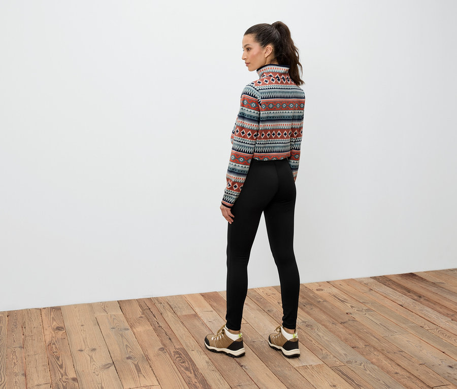 Eine Frau mit braunen Haaren, die zu einem Pferdeschwanz zusammengebunden sind, steht mit dem Rücken zur Kamera und trägt ein gemustertes Langarmshirt, schwarze Leggings und braune Schuhe.