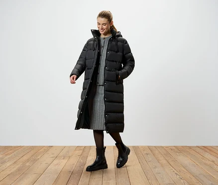 Eine Frau in einer langen, schwarzen Daunenjacke, einem grauen Strickkleid und schwarzen Stiefeln steht auf einem Holzboden.