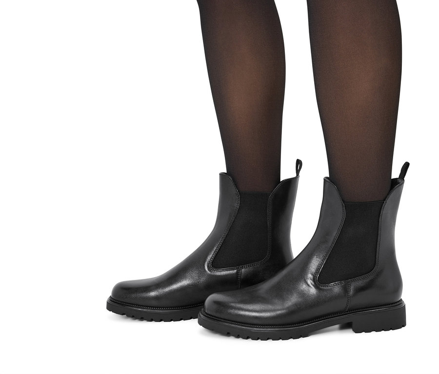 Beine in schwarzen Strumpfhosen, gekleidet in schwarzen Leder-Chelsea-Boots vor einem weißen Hintergrund.