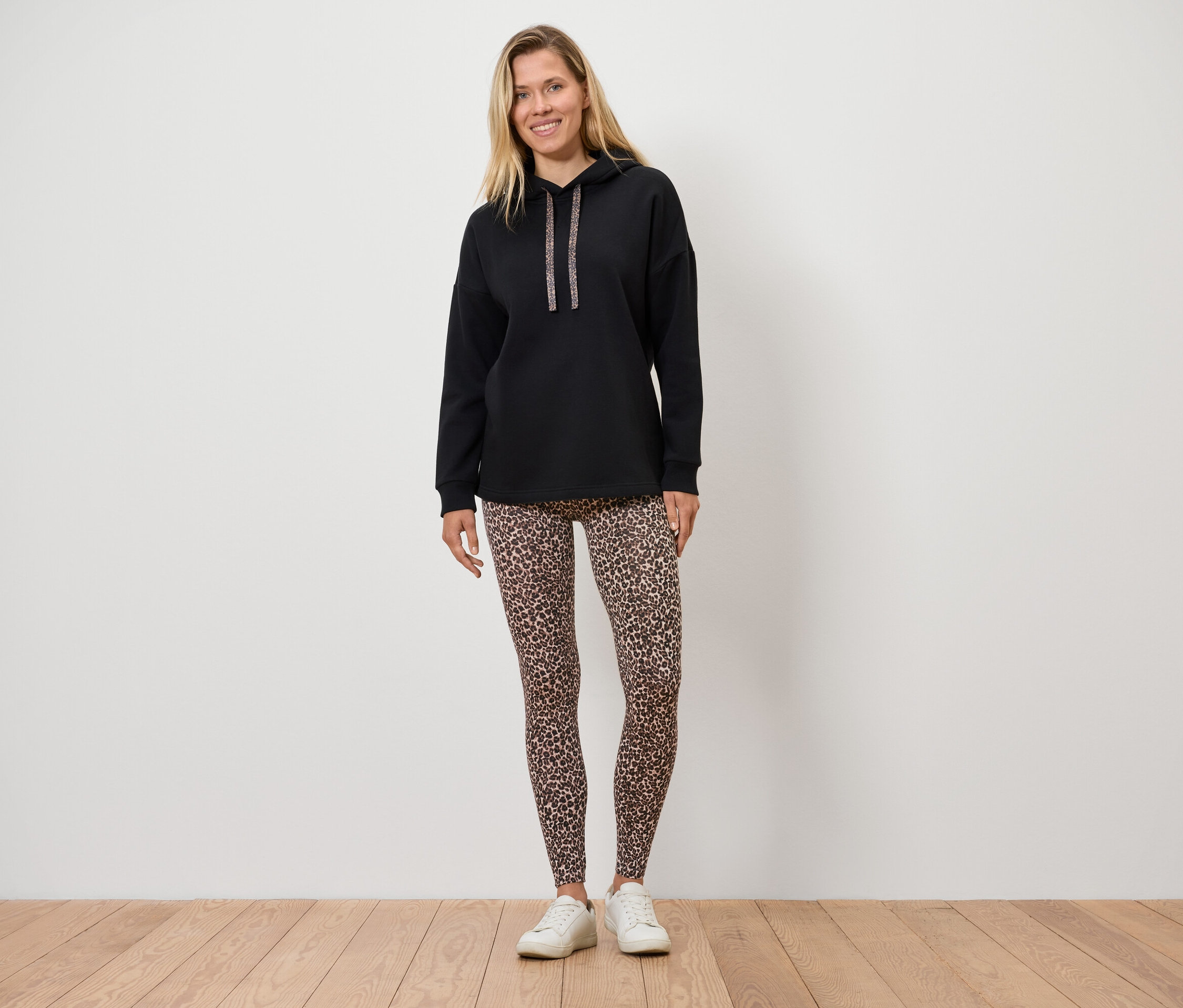 Frau steht auf einem Holzboden und trägt einen schwarzen Hoodie und Leggings mit Tiermuster.