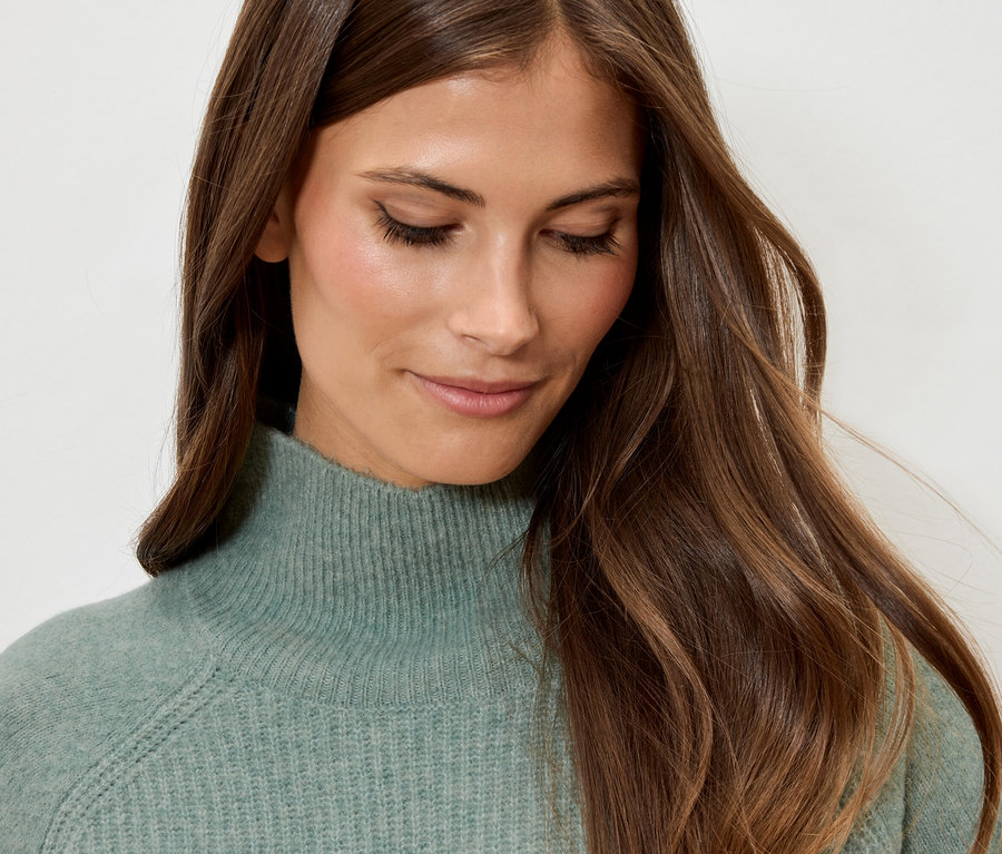 Porträt einer Frau mit braunem, gewelltem Haar, die einen mintgrünen Rollkragenpullover trägt.