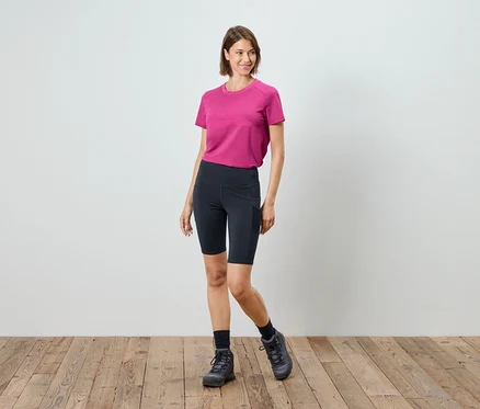 Eine Frau steht auf einem Holzboden und trägt ein pinkfarbenes T-Shirt, schwarze Radlerhosen, Socken und Wanderschuhe.