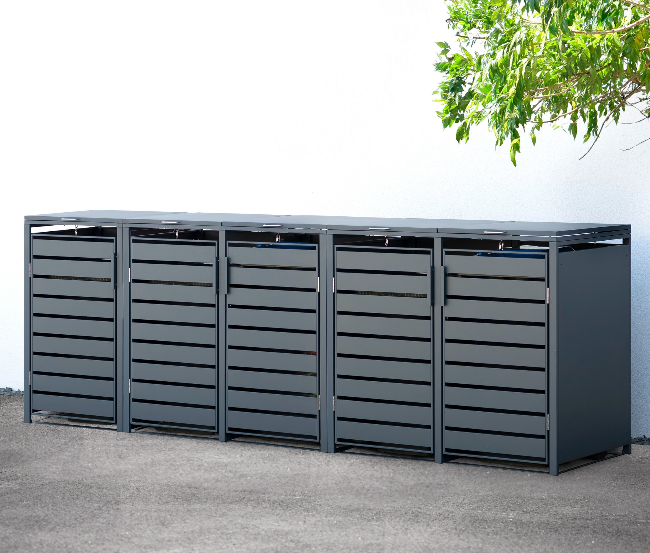 Eine fünfteilige Mülltonnenbox aus Stahl steht draußen an einer weißen Wand.