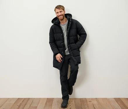 Ein lächelnder Mann mit Bart lehnt an einer Wand und trägt eine schwarze Daunenjacke mit Kapuze, eine schwarze Hose und Stiefel.