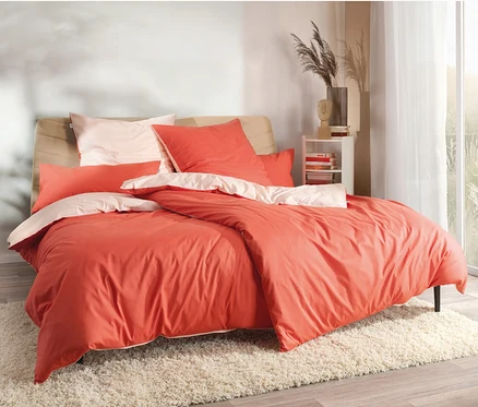 Bett mit korallenroter und pfirsichfarbener SCHÖNER WOHNEN-Kollektion Bettwäsche auf einem weißen, zotteligen Teppich.