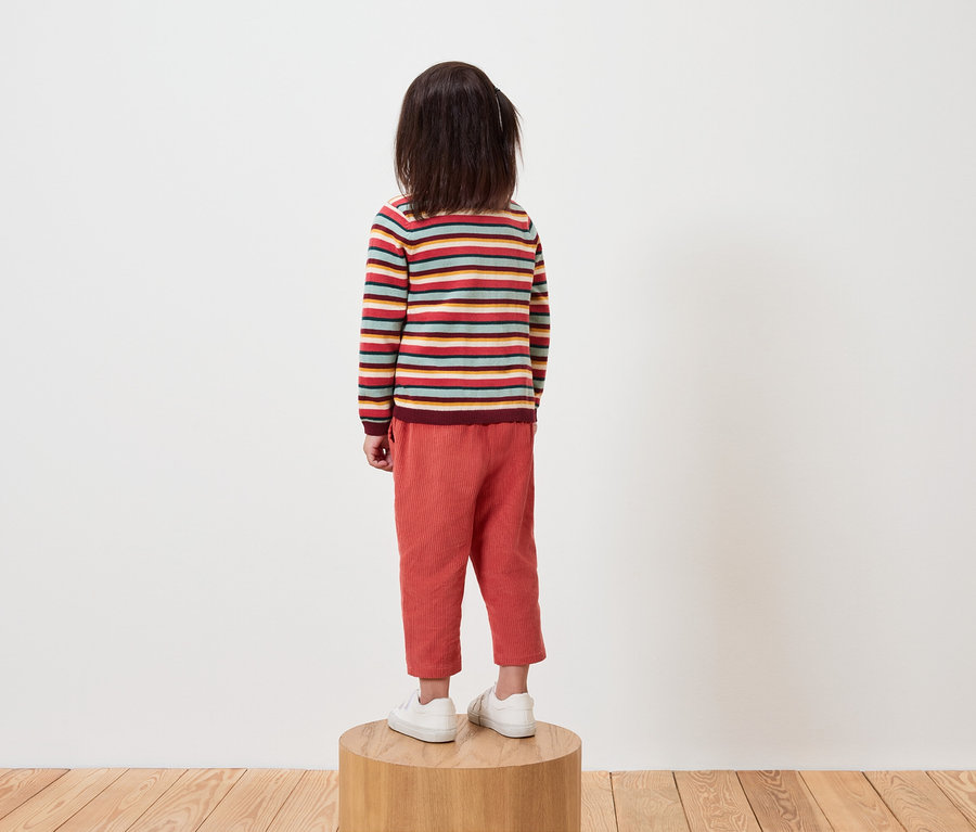 Ein Kind mit dem Rücken zur Kamera steht auf einem Holzsockel und trägt einen gestreiften Pullover und eine orangefarbene Hose.