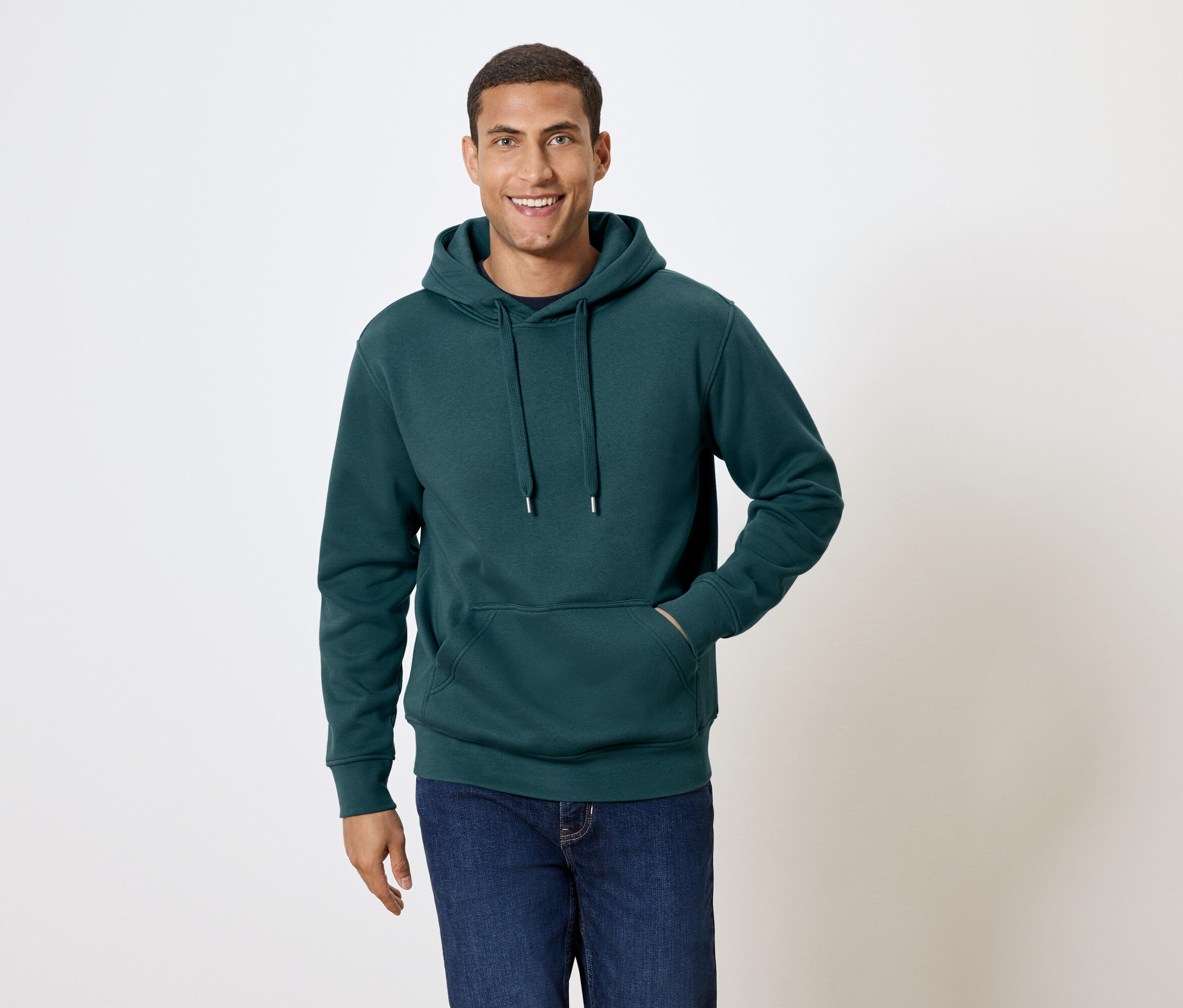 Mann mit kurzen Haaren lächelt und posiert in einem dunkelgrünen Hoodie und blauen Jeans.