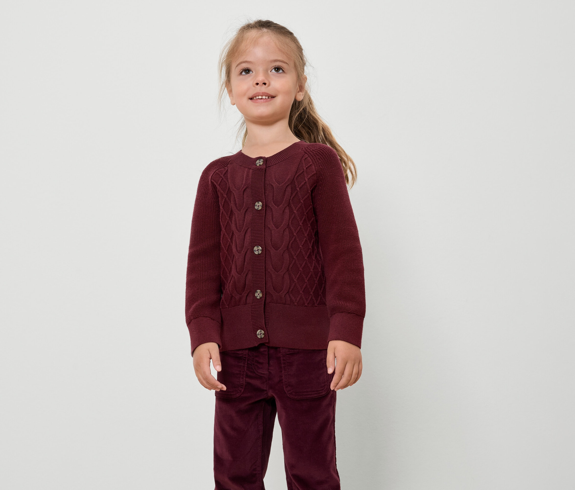 Mädchen in bordeauxrotem Pullover und Hose steht vor einem weißen Hintergrund.