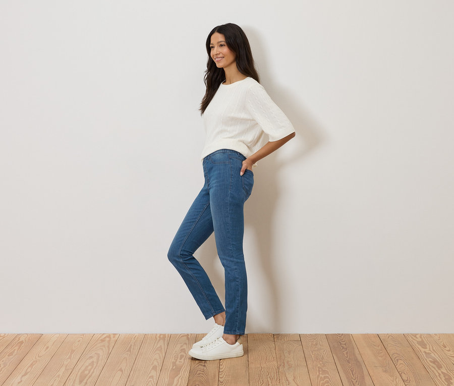 Frau steht seitlich vor einer weißen Wand, trägt blaue Jeans, einen weißen Pullover und weiße Turnschuhe.