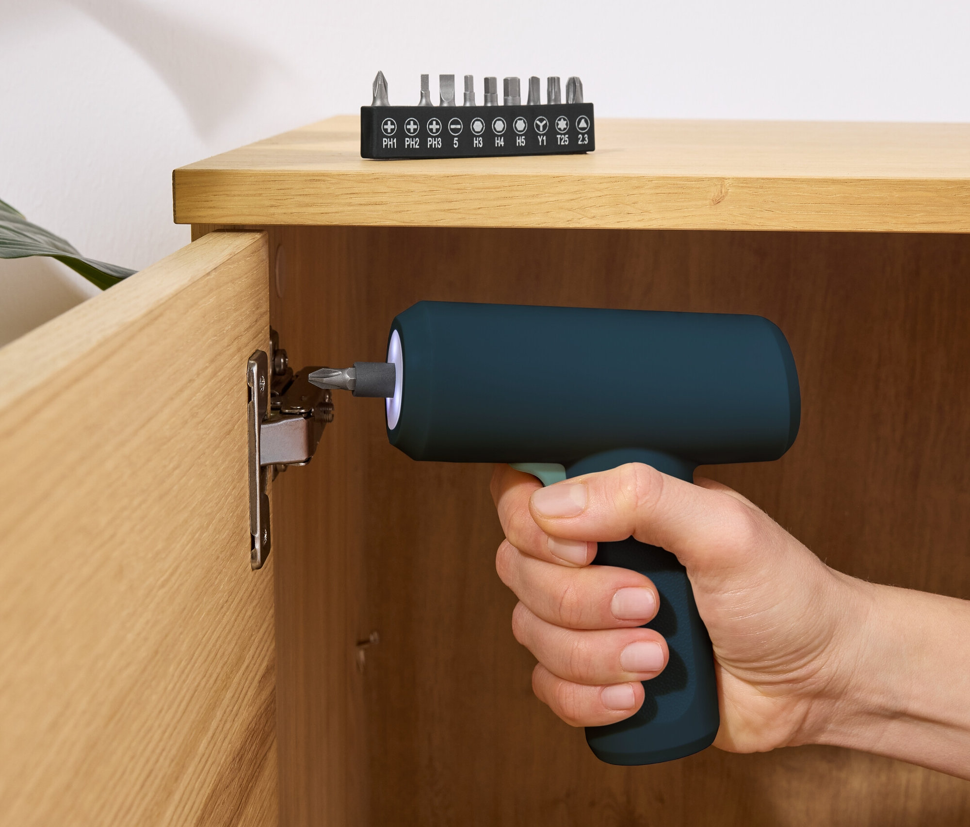 Eine Hand hält einen blauen Mini-Akkuschrauber 3,6 V und schraubt eine Schraube in einen Holzschrank. Ein Satz Bits liegt auf der Oberseite des Schranks.