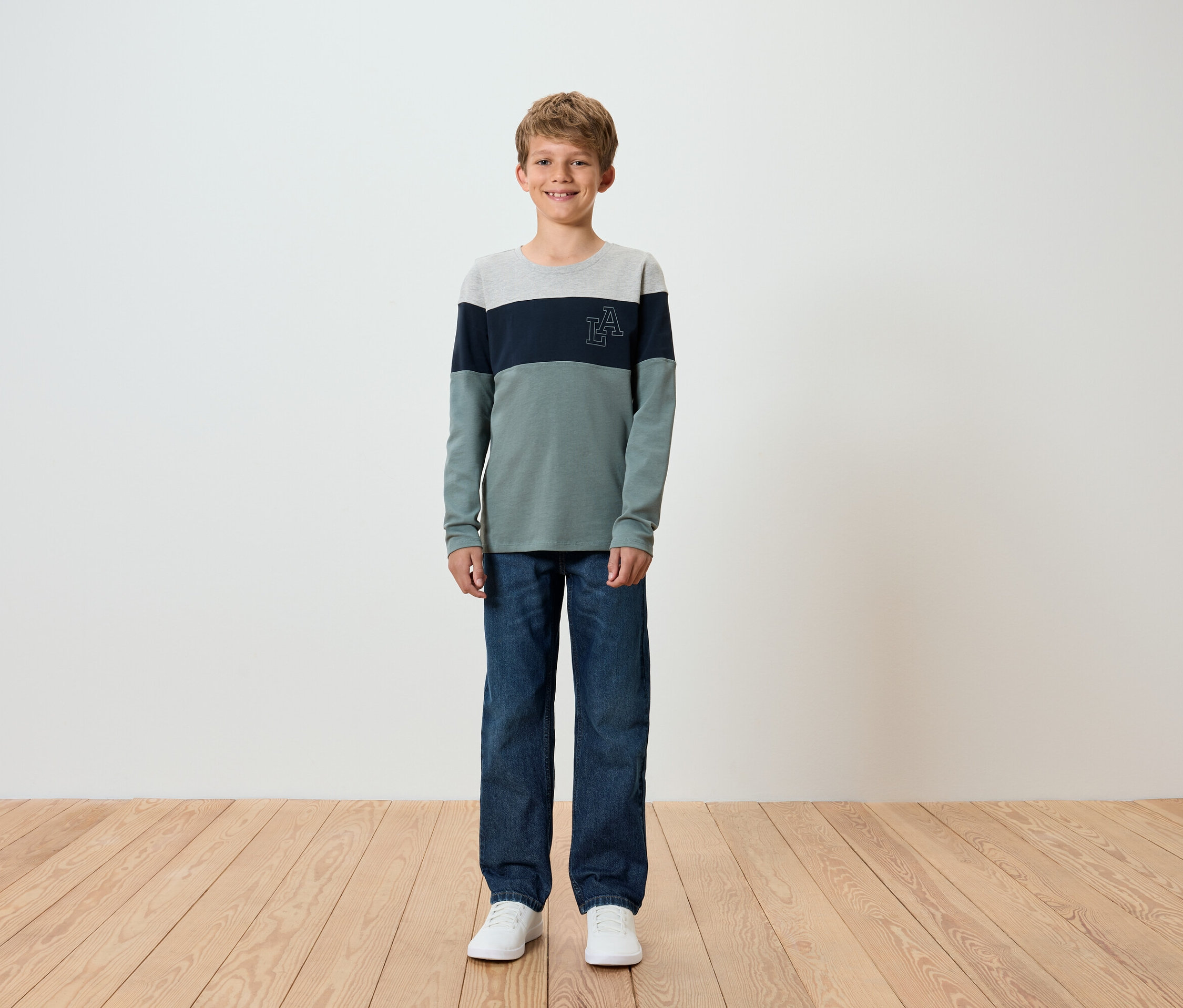 Junge steht auf Holzboden, trägt blaue Jeans und ein langärmliges T-Shirt.