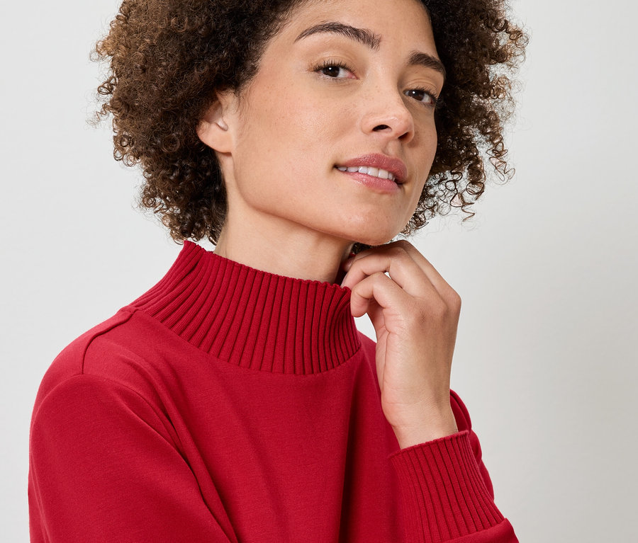 Detailporträt einer Frau mit lockigem Haar, die einen roten Rollkragenpullover trägt und ihre Hand am Hals hat.