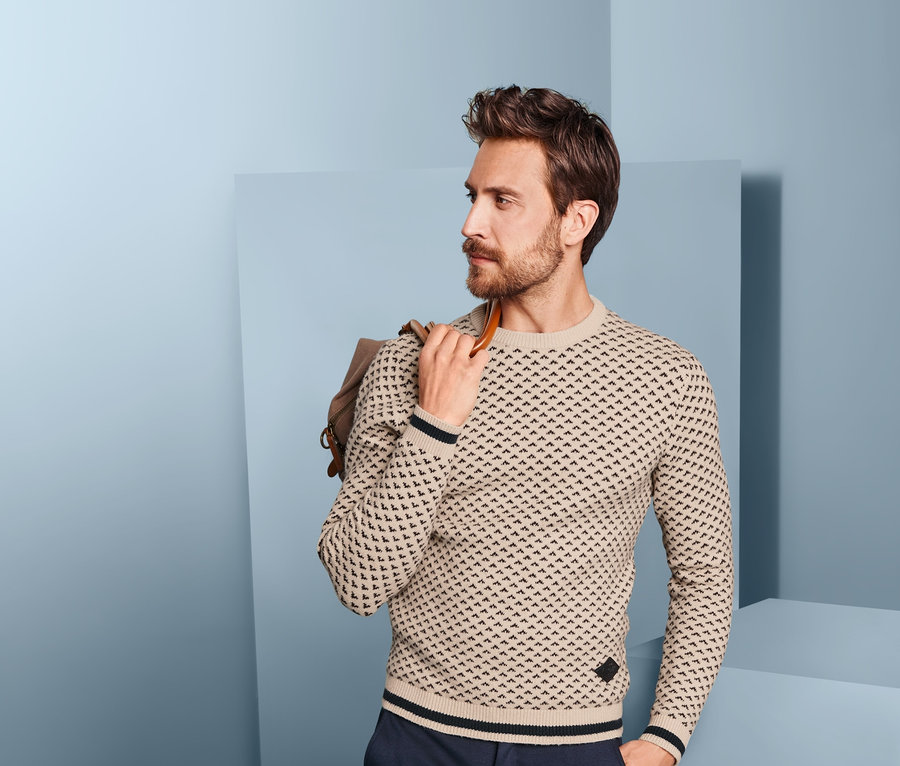 Mann mit Bart trägt einen Strickpullover und dunkelblaue Hosen. Er hat eine Tasche über der Schulter.