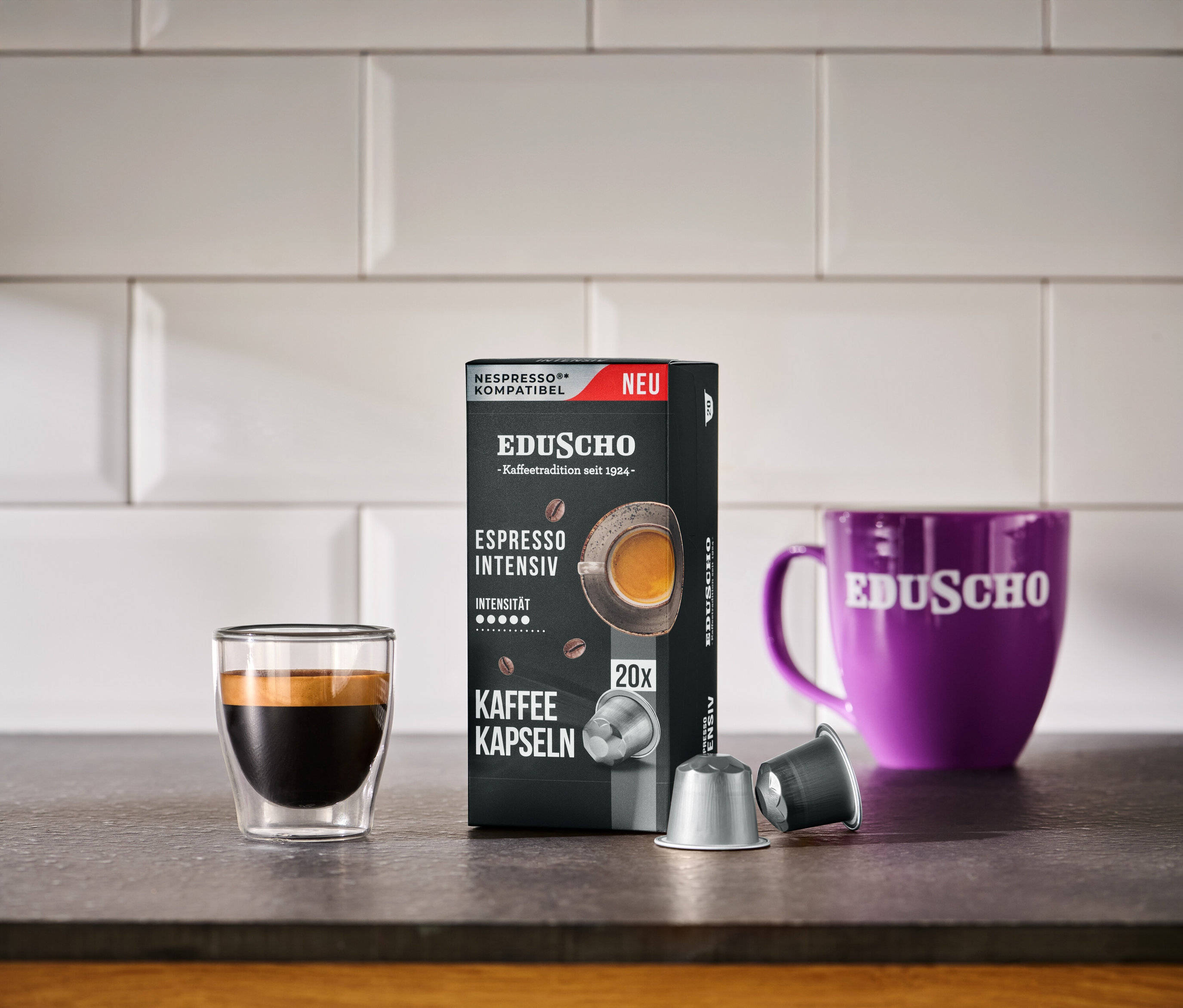 Eine schwarze Packung EDUSCHO Espresso Intensiv neben einem Espressoglas, einer lila Tasse und zwei Kapseln.