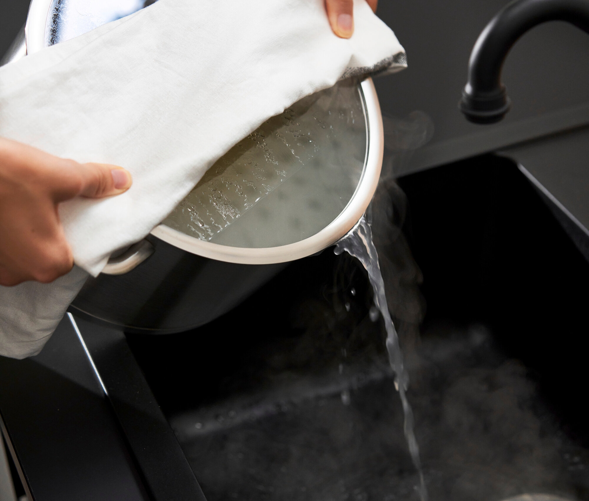 Person hält Topf mit Wasser über schwarzem Spülbecken, Wasser läuft aus dem Topf.