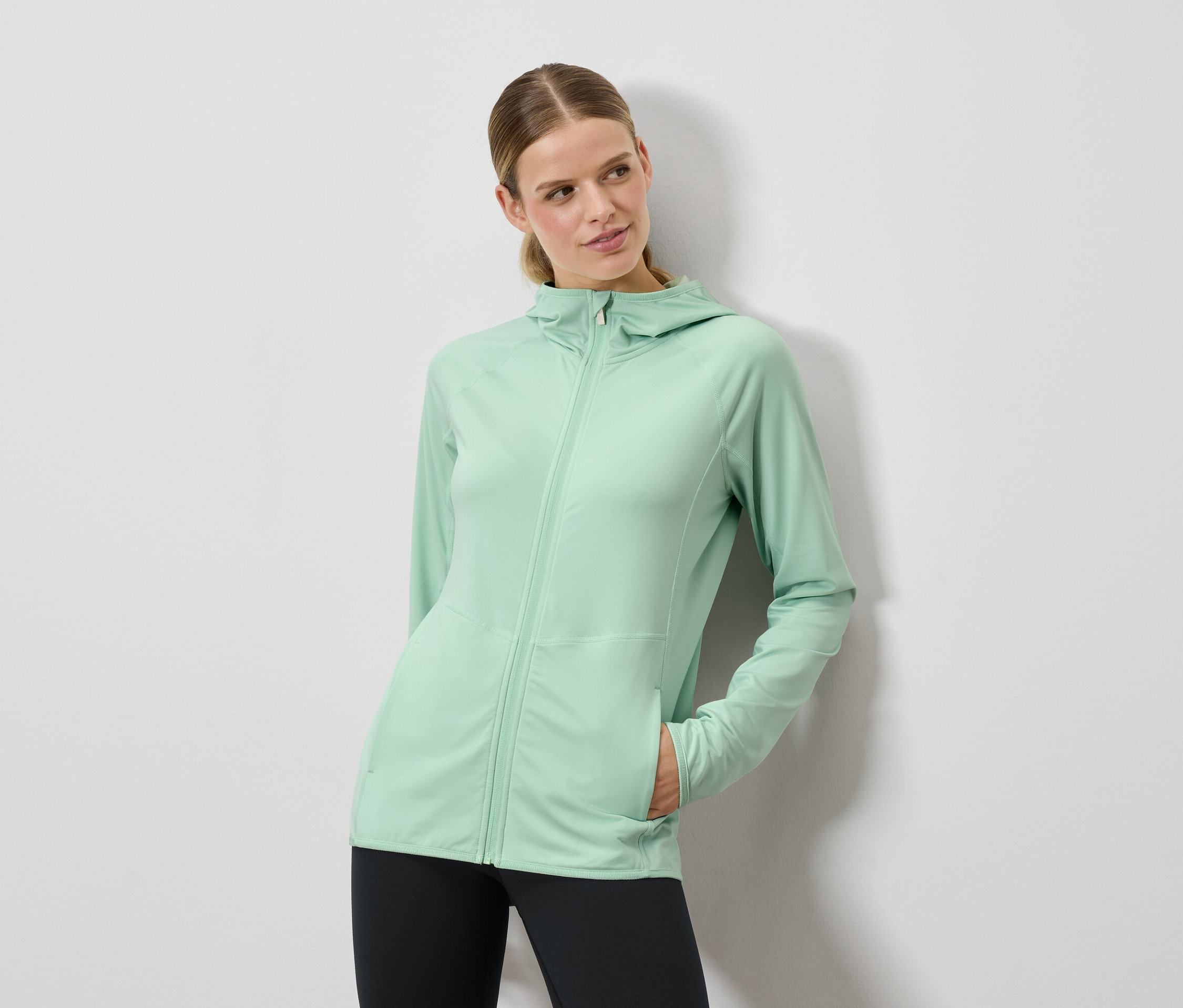 Frau posiert in mintgrüner Kapuzenjacke und schwarzen Leggings.