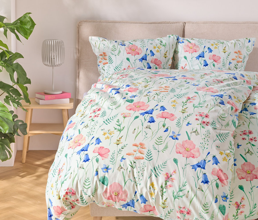 Bett mit floraler Bettwäsche, daneben ein Tisch mit Lampe und Büchern, links eine Pflanze.