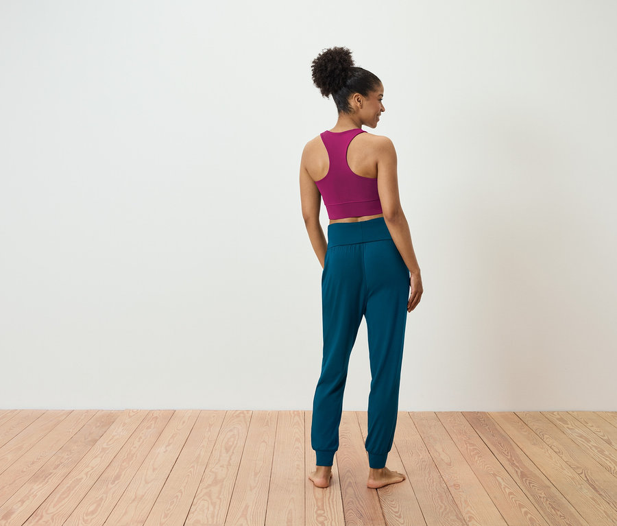 Eine Frau steht mit dem Rücken zur Kamera und trägt einen magentafarbenen Sport-BH und blaugrüne Jogginghose.